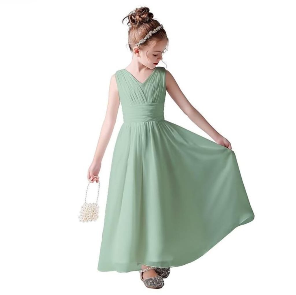 A young girl wearing a sleeveless, mint green chiffon dress with a pleated bodice and a flowing skirt. She is holding the skirt with one hand and has her hair styled in a bun with a floral headband.