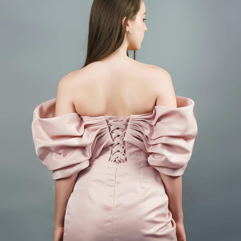 A woman wearing an elegant, pastel pink satin chiffon dress with voluminous puffed sleeves and a fitted bodice featuring a lace-up back detail, standing against a neutral background.