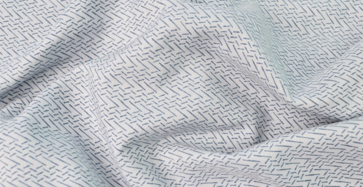 Close-up image of light-colored Sea Island cotton fabric with fine, small, dark dashed lines forming a subtle, intricate pattern.
