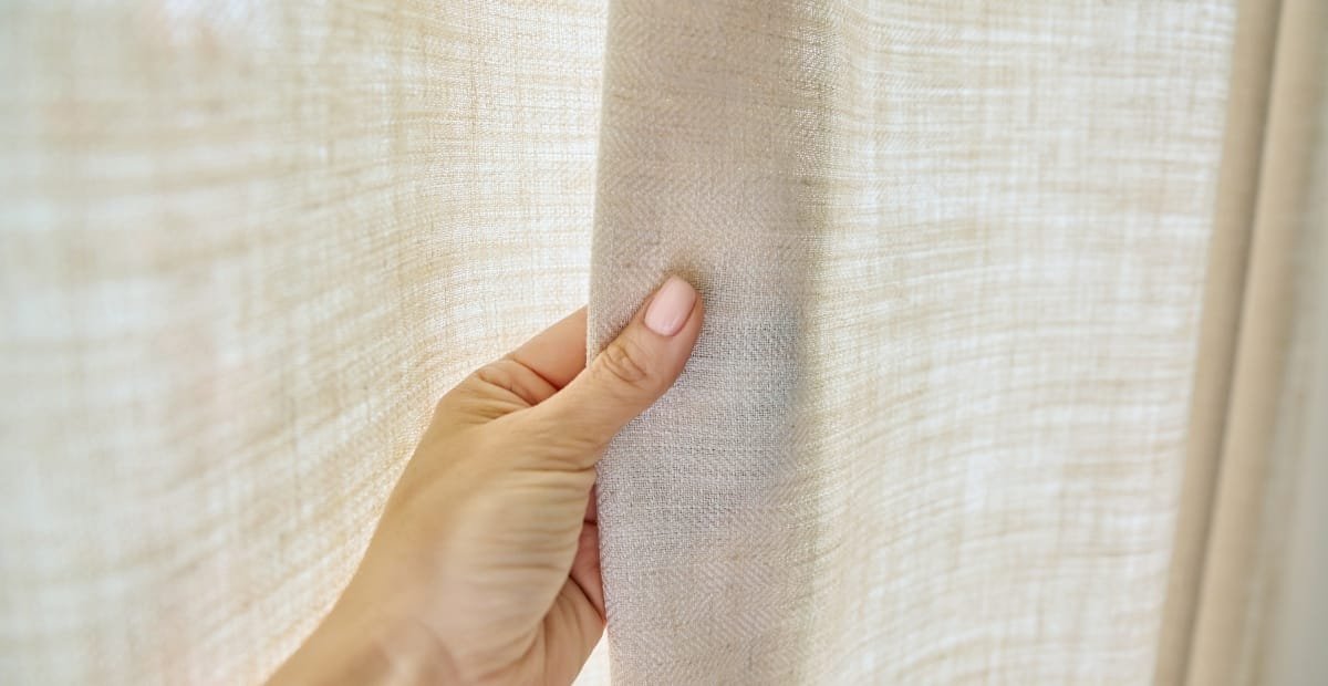 A hand gently holding back a light-colored fabric curtain, revealing more of the same material.
