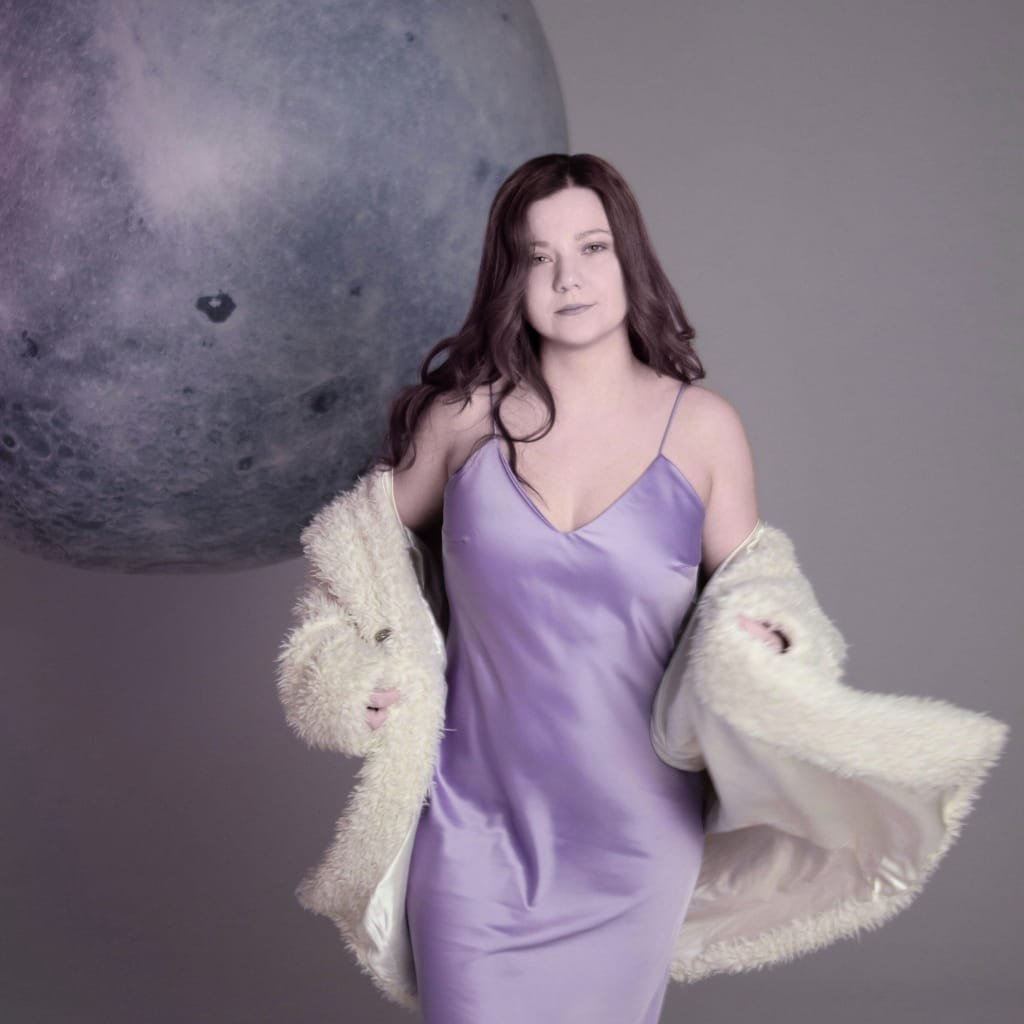A woman in a lavender satin slip dress with thin straps, draped in a luxurious cream-colored fur coat, standing against a plain gray background with a large moon-like sphere floating behind her.
