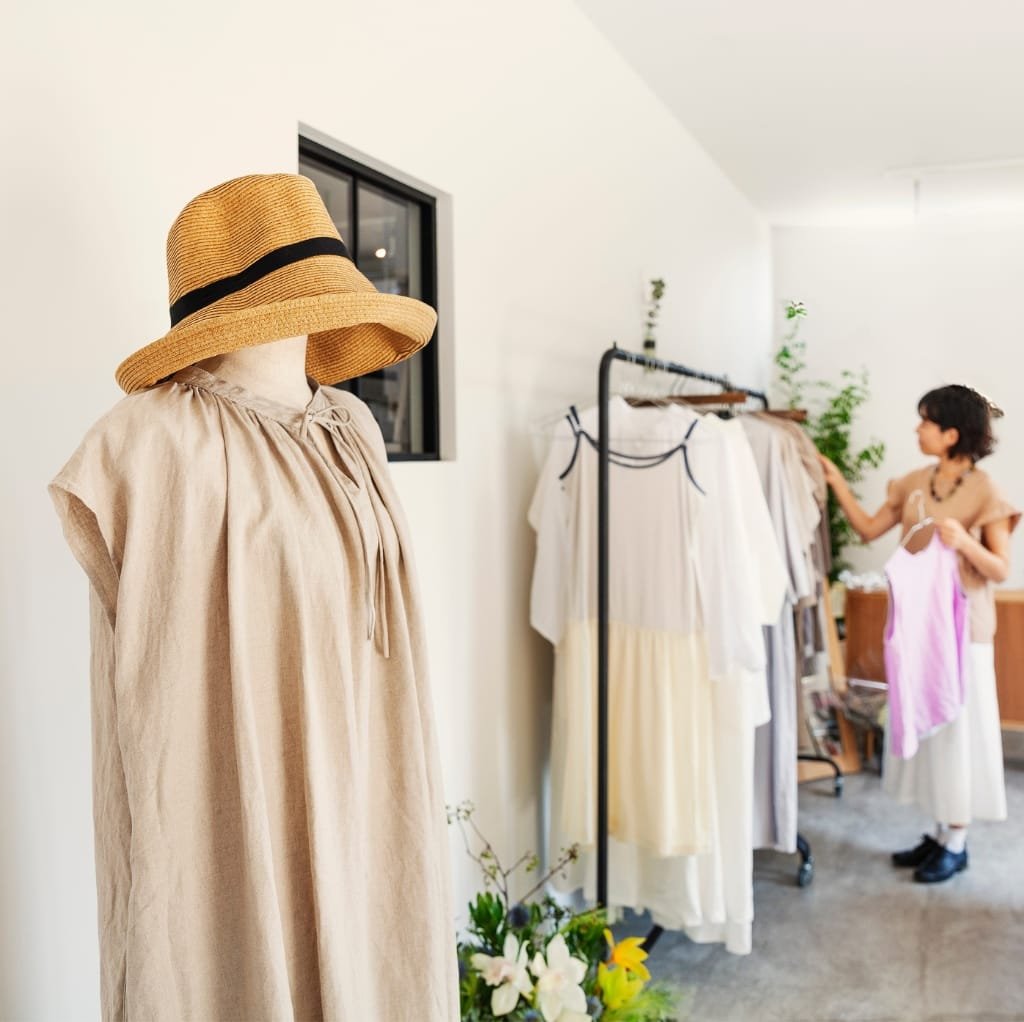 A beige smock cotton dress on a mannequin wearing a wide-brimmed straw hat, displayed in a bright, minimalistic boutique or show space. In the background, a woman with short dark hair is browsing through hanging garments on a clothing rack.