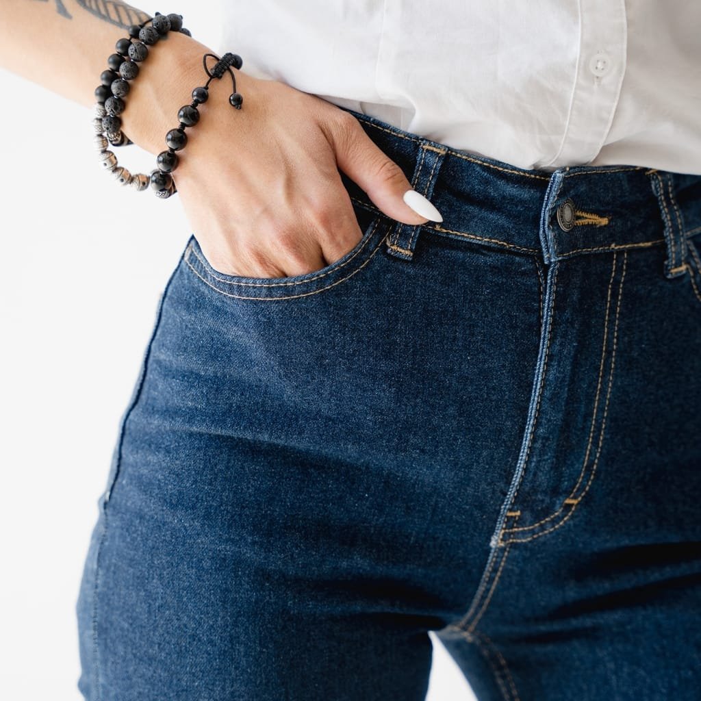 Close-up of a person wearing dark blue, high-waisted stretch denim jeans with their hand tucked into the front pocket. The person has a white shirt, and is accessorizing with black beaded bracelets on their wrist.
