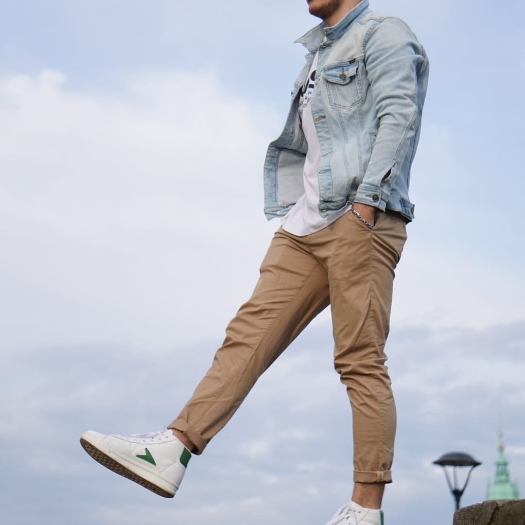 A person standing outdoors against a cloudy sky, wearing a light chambray denim shirt layered over a T-shirt, paired with beige chinos, white sneakers with green accents, and a silver bracelet. One leg is raised, giving a casual, stylish pose.