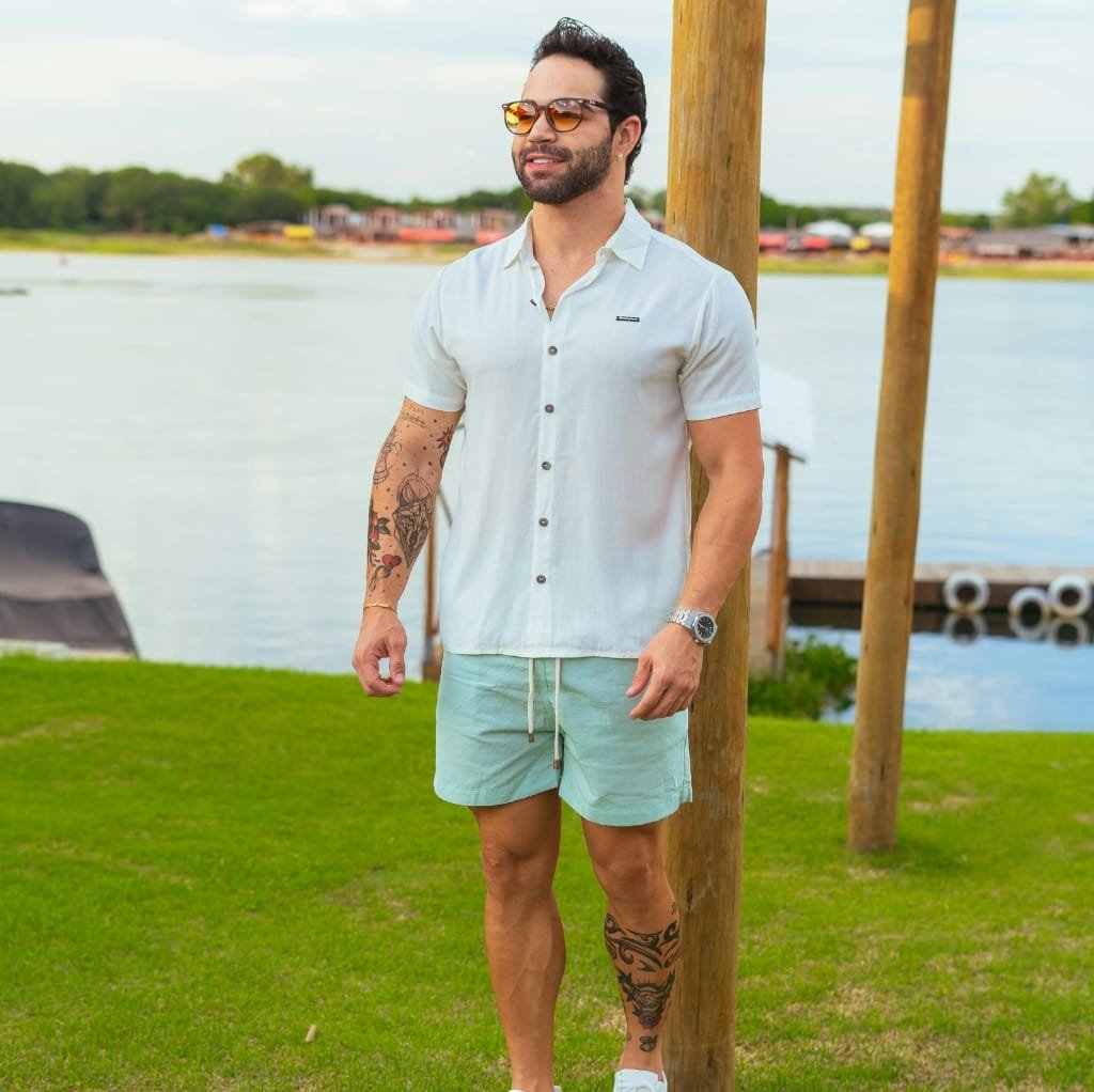 A man standing outdoors near a body of water, wearing a white linen short-sleeve shirt, light-colored shorts, sunglasses, and a wristwatch.