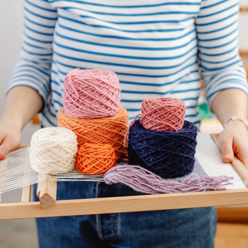 A person holding a wooden tray with several colorful balls of yarn in various sizes, including pink, orange, navy blue, and white, with a piece of purple yarn lying on the tray. The person is wearing a blue and white striped shirt and blue jeans.