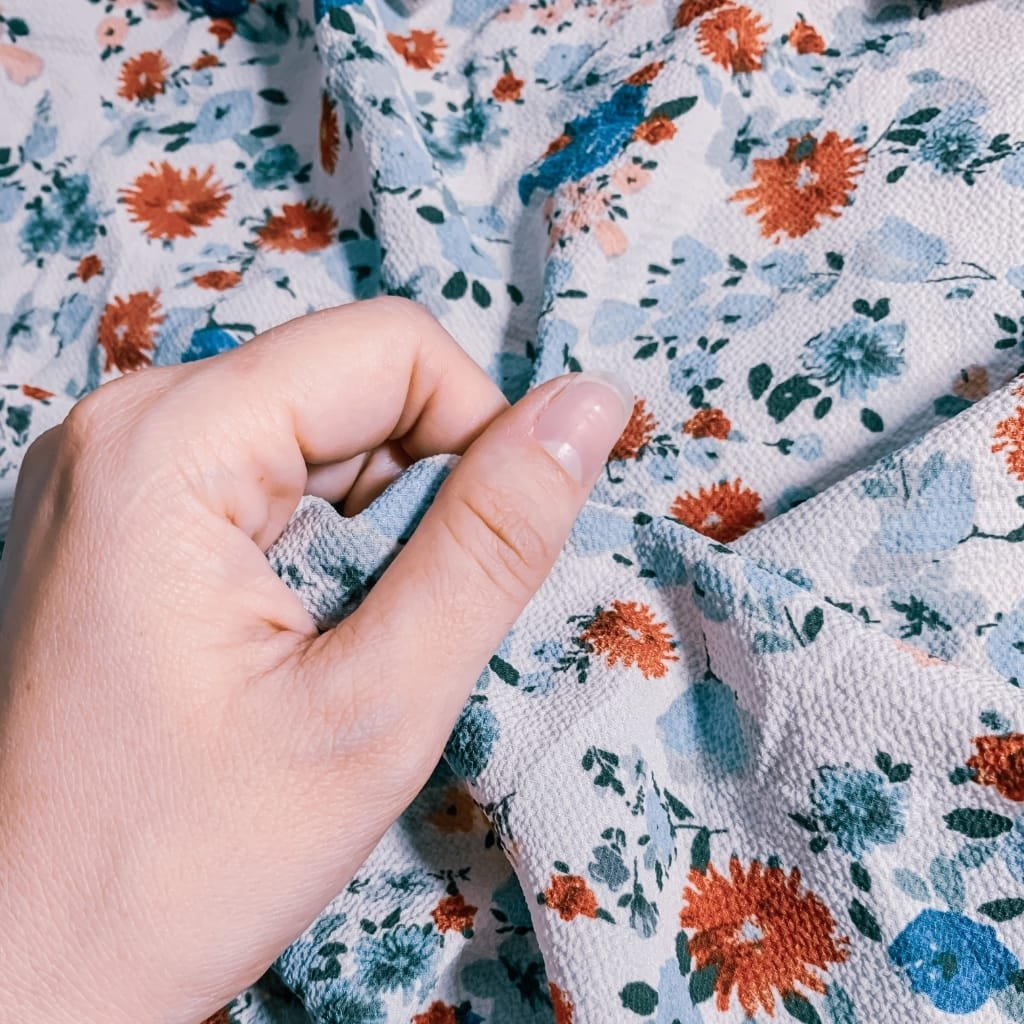 Close-up of a person's hand holding the corner of a floral printed chiffon fabric, showing the texture and pattern.