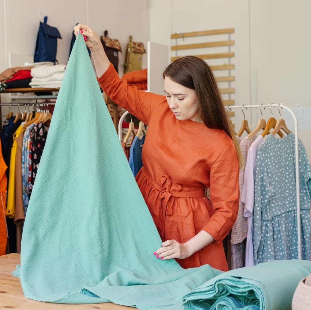 A woman in an orange dress is inspecting a large piece of light blue thick cotton fabric in a fabric or material store. There are other fabrics and clothing items displayed in the background.