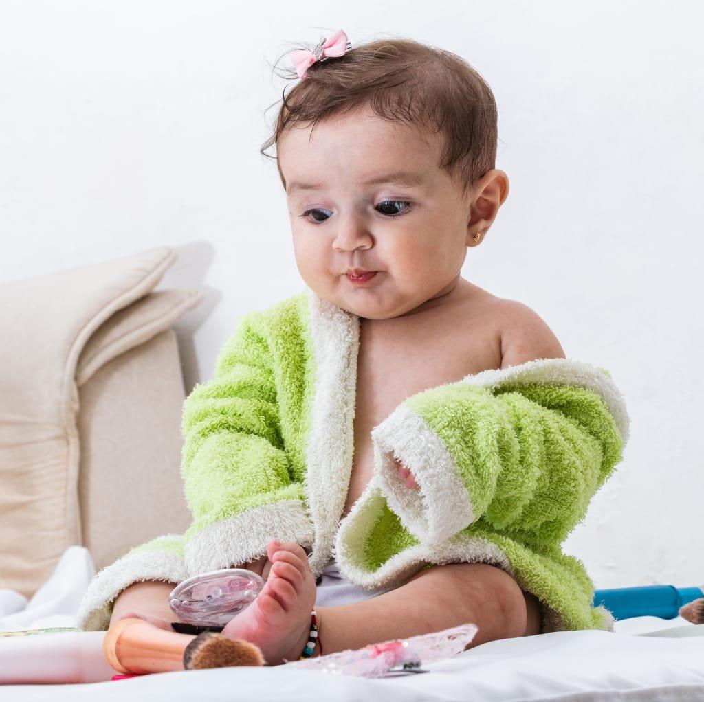 A young child with light brown hair, wearing a green and white fluffy towel or robe, sitting on a bed and looking thoughtfully at their foot with medical devices attached.