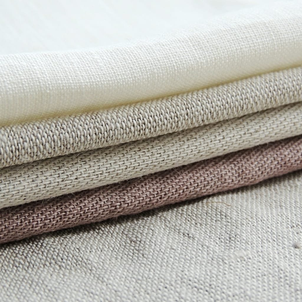 A close-up shot of stacked, folded fabrics in neutral shades, showcasing textured weaves and natural finishes.