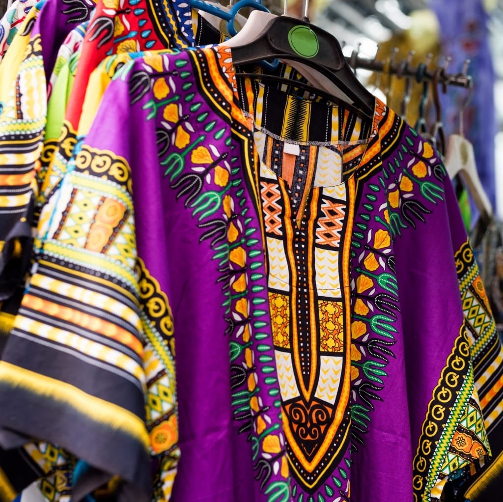 Colorful, traditional cotton kaftan dresses with intricate patterns hanging on a rack at a market. The dresses feature vibrant purple, yellow, orange, and green designs with detailed embroidery and geometric motifs.