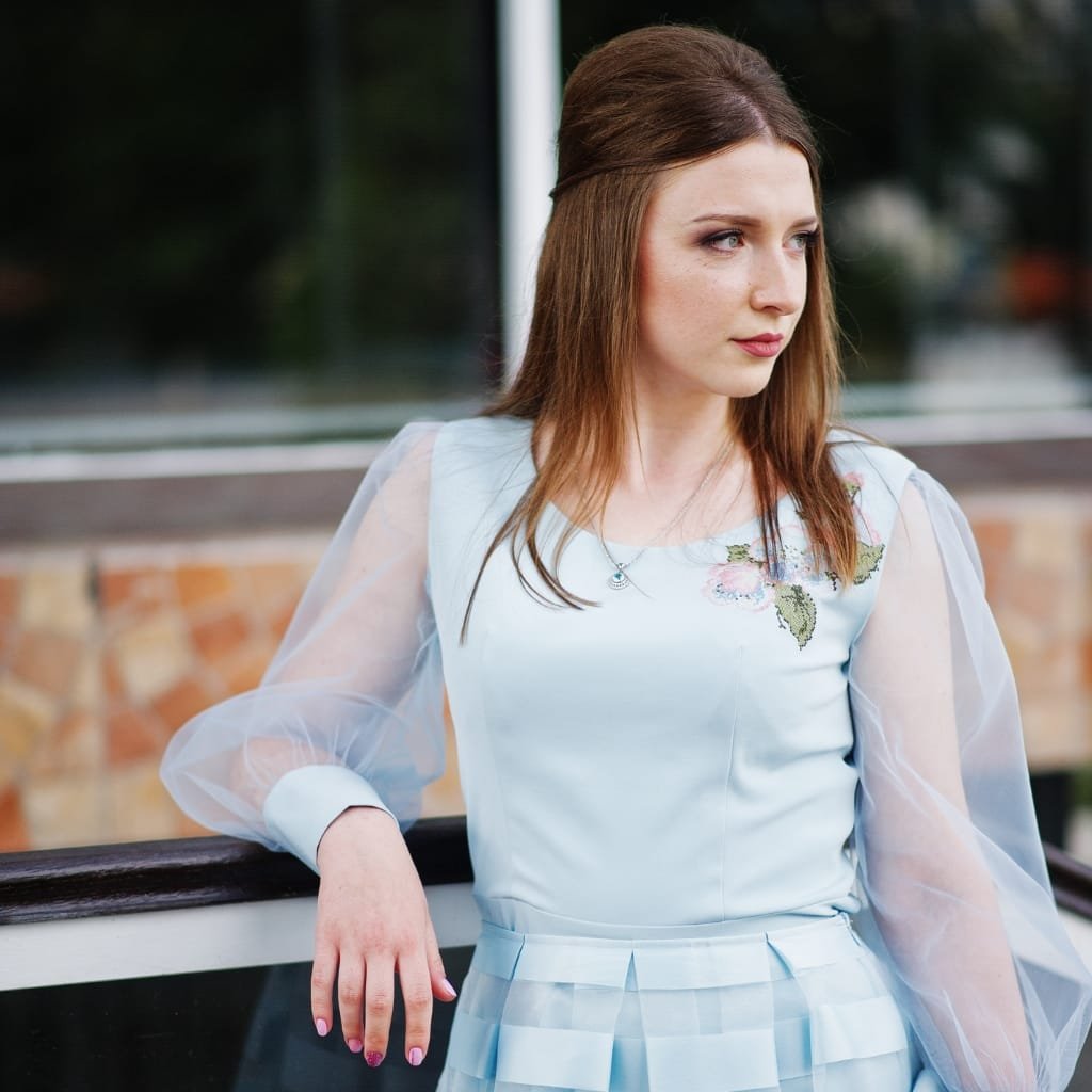 A woman with light skin, long reddish-brown hair, and wearing a light blue dress with sheer, puffy sleeves stands outdoors near a railing, looking to her right.