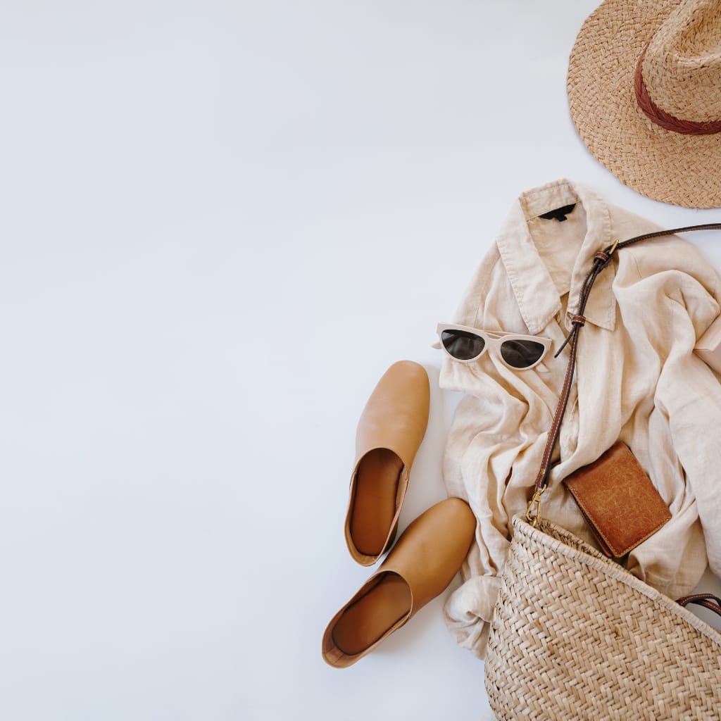 A flat lay of summer travel essentials, including a wide-brimmed straw hat, beige linen shirt, brown slip-on shoes, a pair of black sunglasses, a small brown wallet, a woven straw bag, and a small bottle, all arranged on a white background.