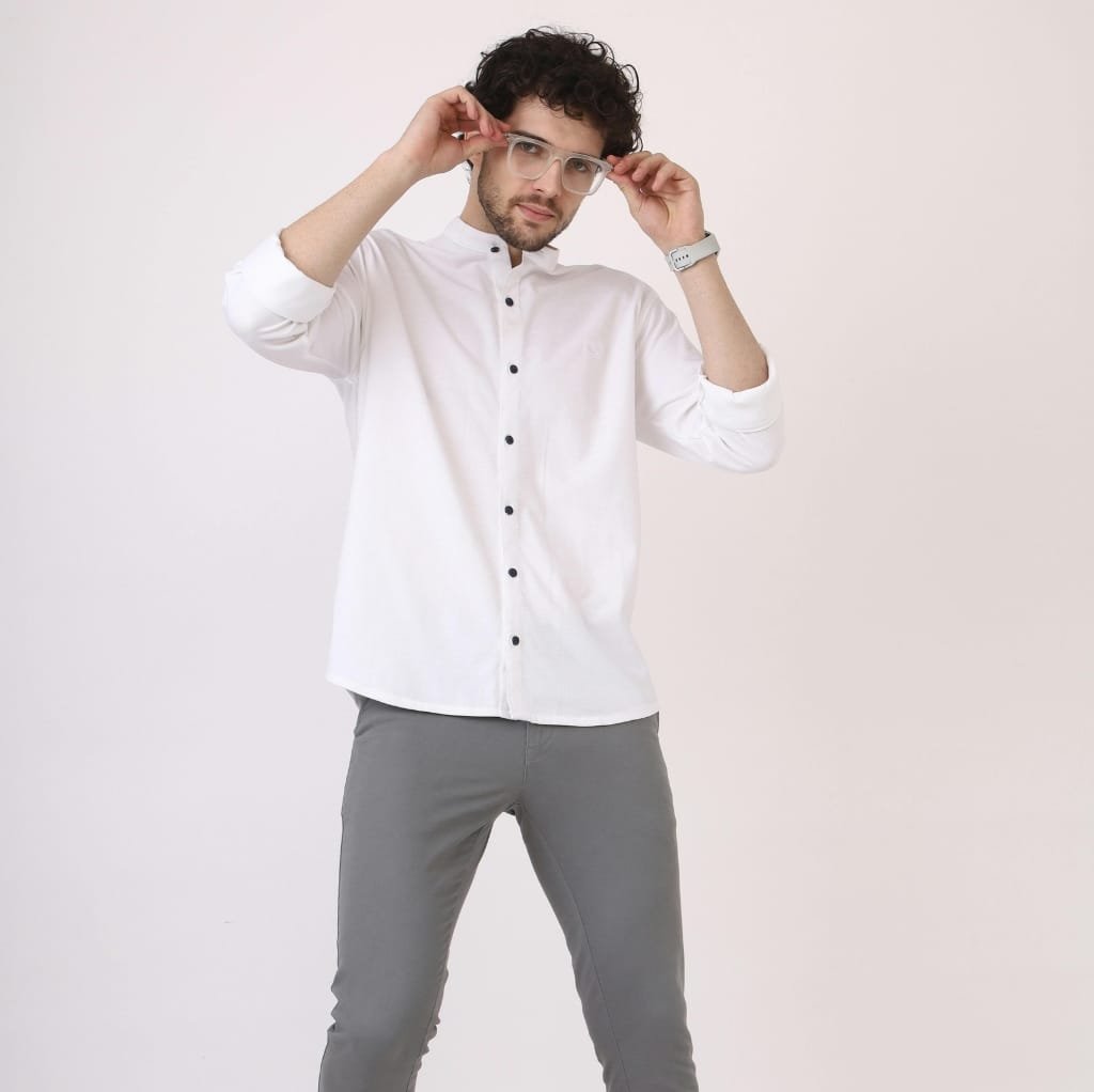 A man with curly dark hair and glasses, wearing a white button-up shirt and gray pants, adjusting his glasses against a plain background, illustrating a smart-casual style.