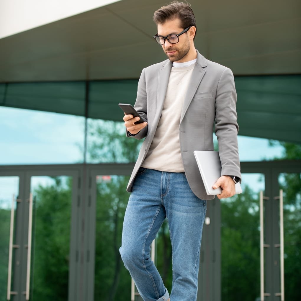 A man dressed in a light-colored polo shirt and a gray sport coat, holding a laptop in one hand and a smartphone in the other, walking outdoors.