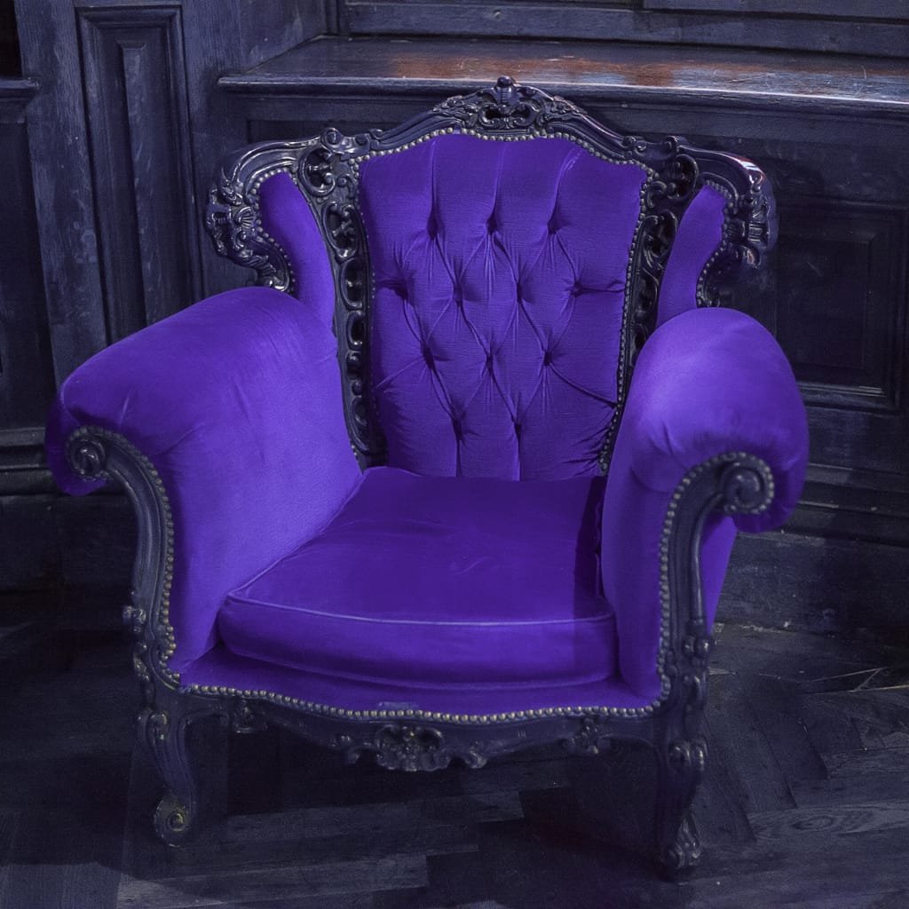 A vintage-style armchair with ornate wooden carvings and deep purple velvet upholstery, set against a dark wooden wall.