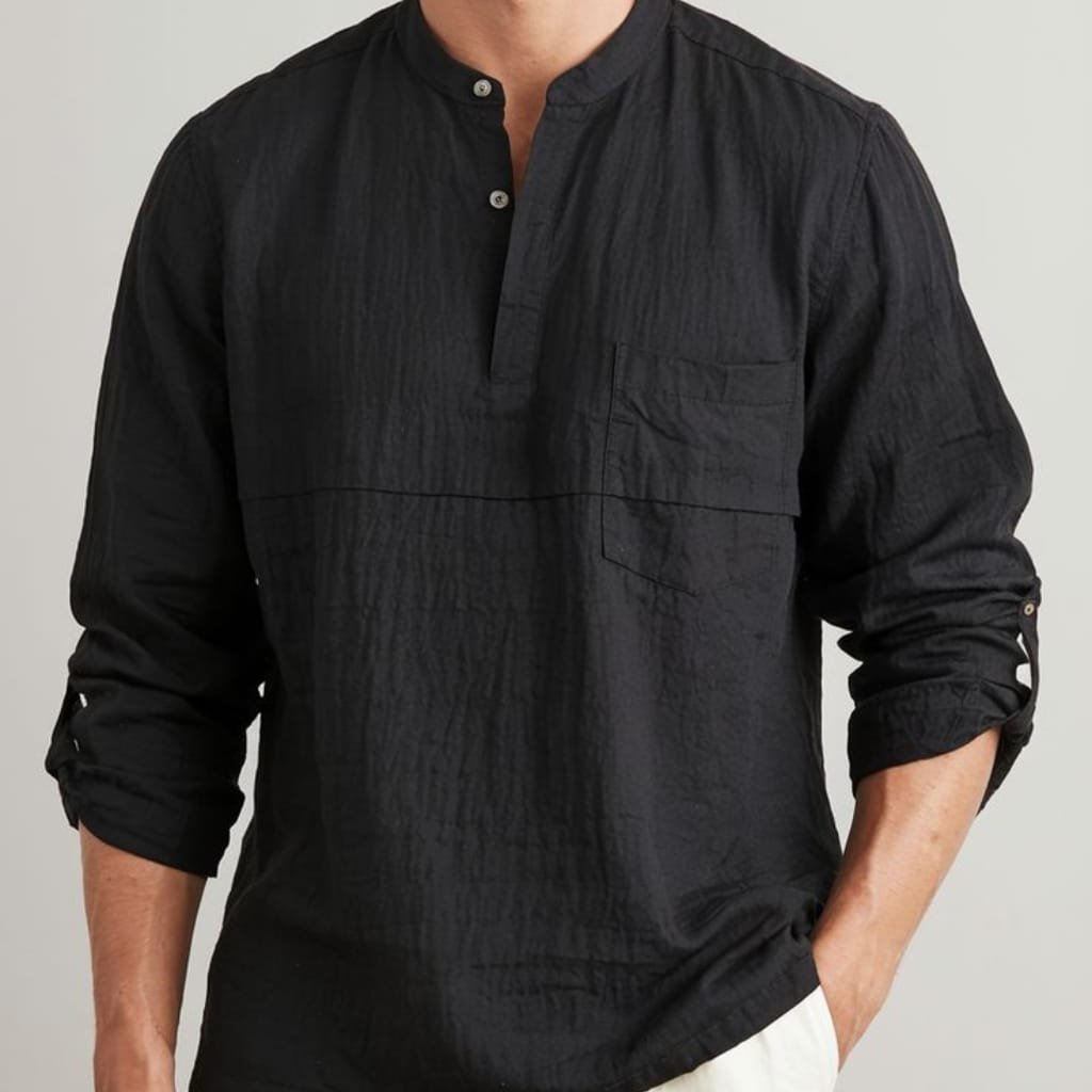 A person wearing a black, long-sleeve, button-up shirt with a stand collar, displayed with sleeves rolled up and their left hand in their pocket. The shirt has a textured, slightly crinkled fabric with a chest pocket visible on the left side.