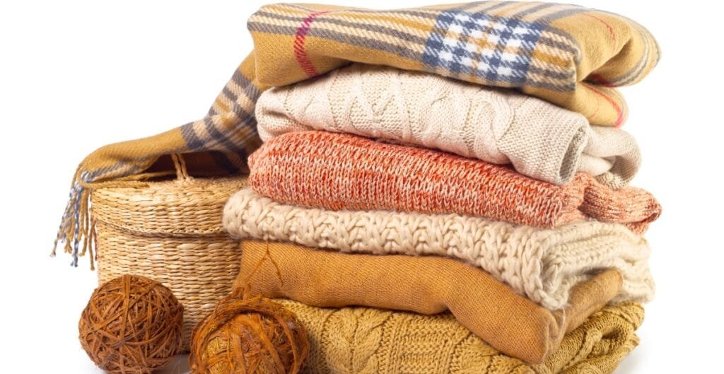 A neatly stacked pile of folded sweaters and scarves next to woven baskets and balls of yarn.