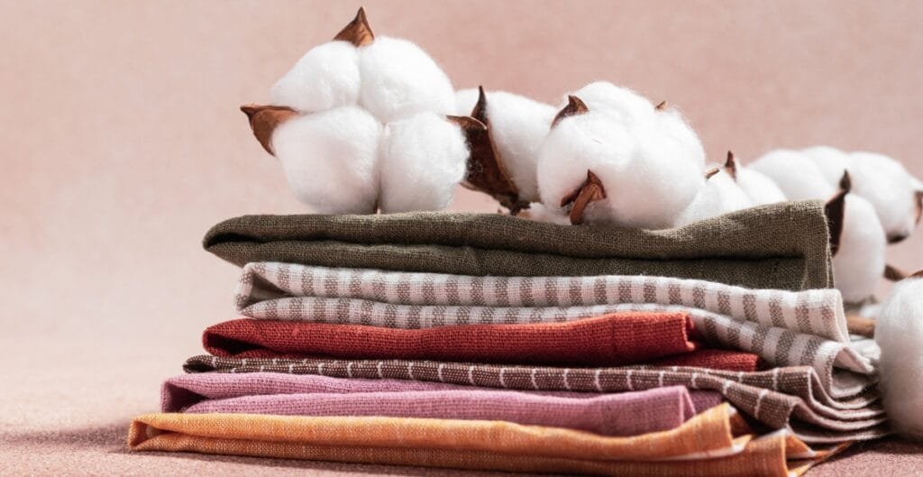 A stack of folded cloth napkins in various colors and patterns, placed on a surface with a bunch of cotton plants resting behind them.