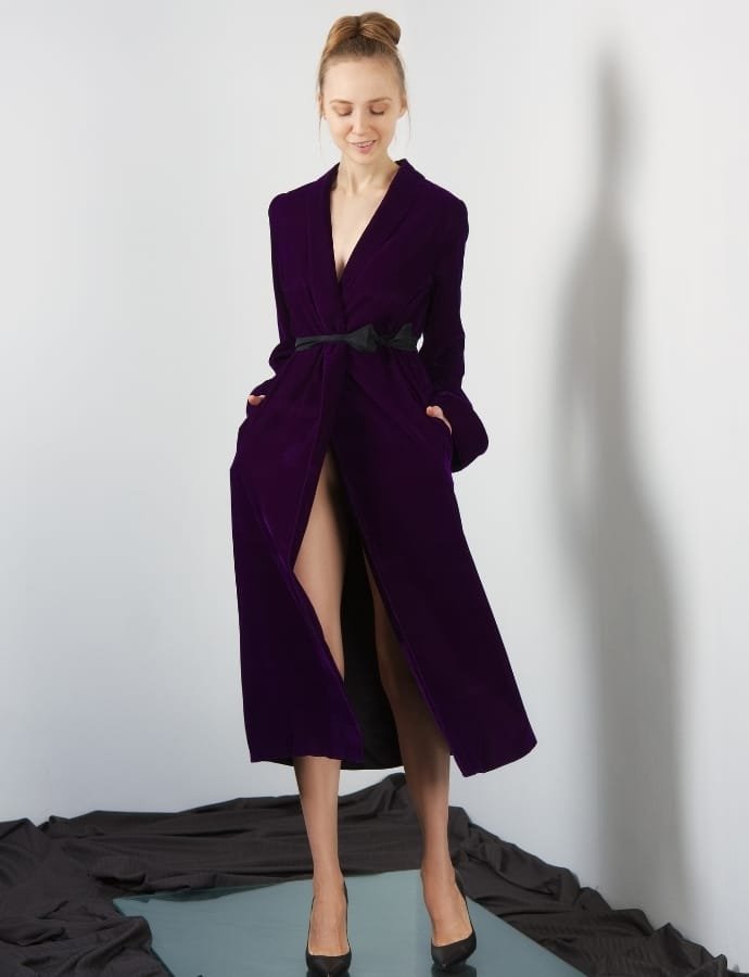 A woman standing against a white wall, wearing a long, deep purple velour coat with a black belt, black high heels, and a subtle smile, with her hands in her pockets.