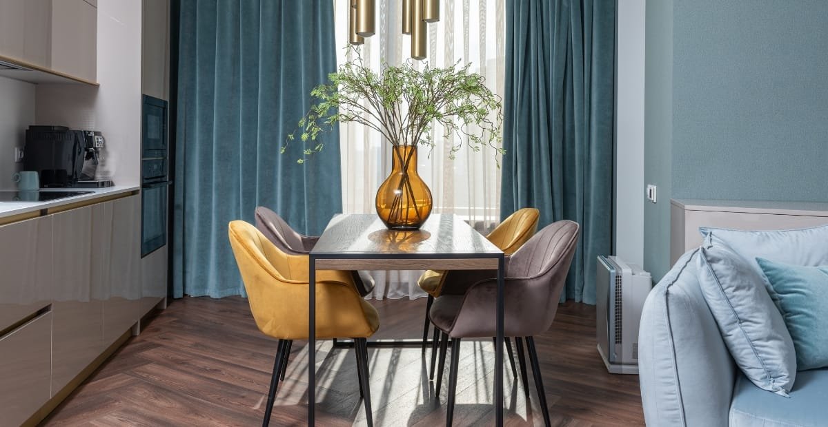 Close-up of a modern home interior featuring a dining table with a large amber glass vase containing a leafy green plant, surrounded by colorful velvet upholstered chairs, with curtains and a cozy sofa in the background.