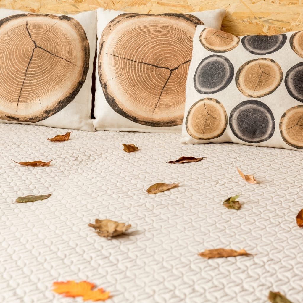 Decorative pillows with images of tree rings depicting cross-sections of wood, arranged on a bed with a textured white bedspread, scattered with autumn leaves, and a wooden wall in the background.