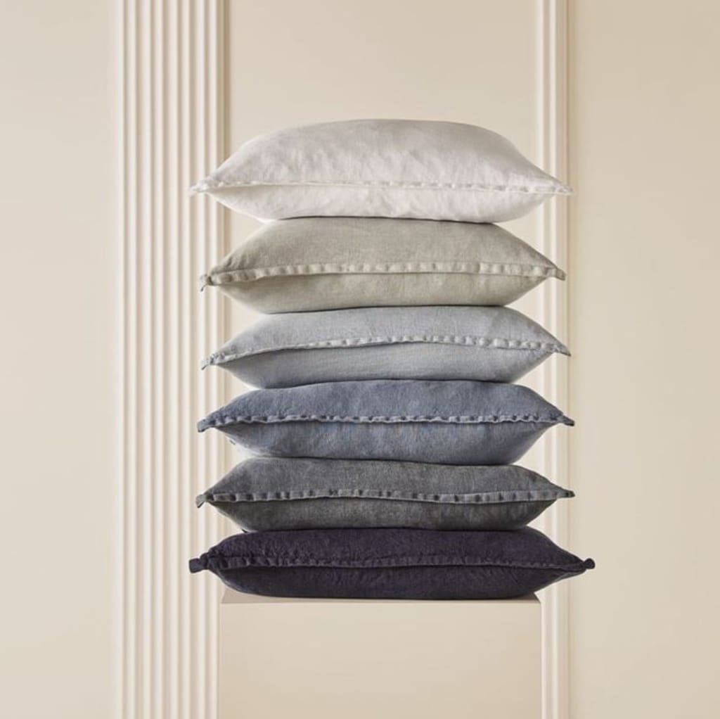 A neatly stacked pyramid of six plush, multi-colored cushions in shades of white, beige, gray, and black, set against a neutral background with vertical wall accents.