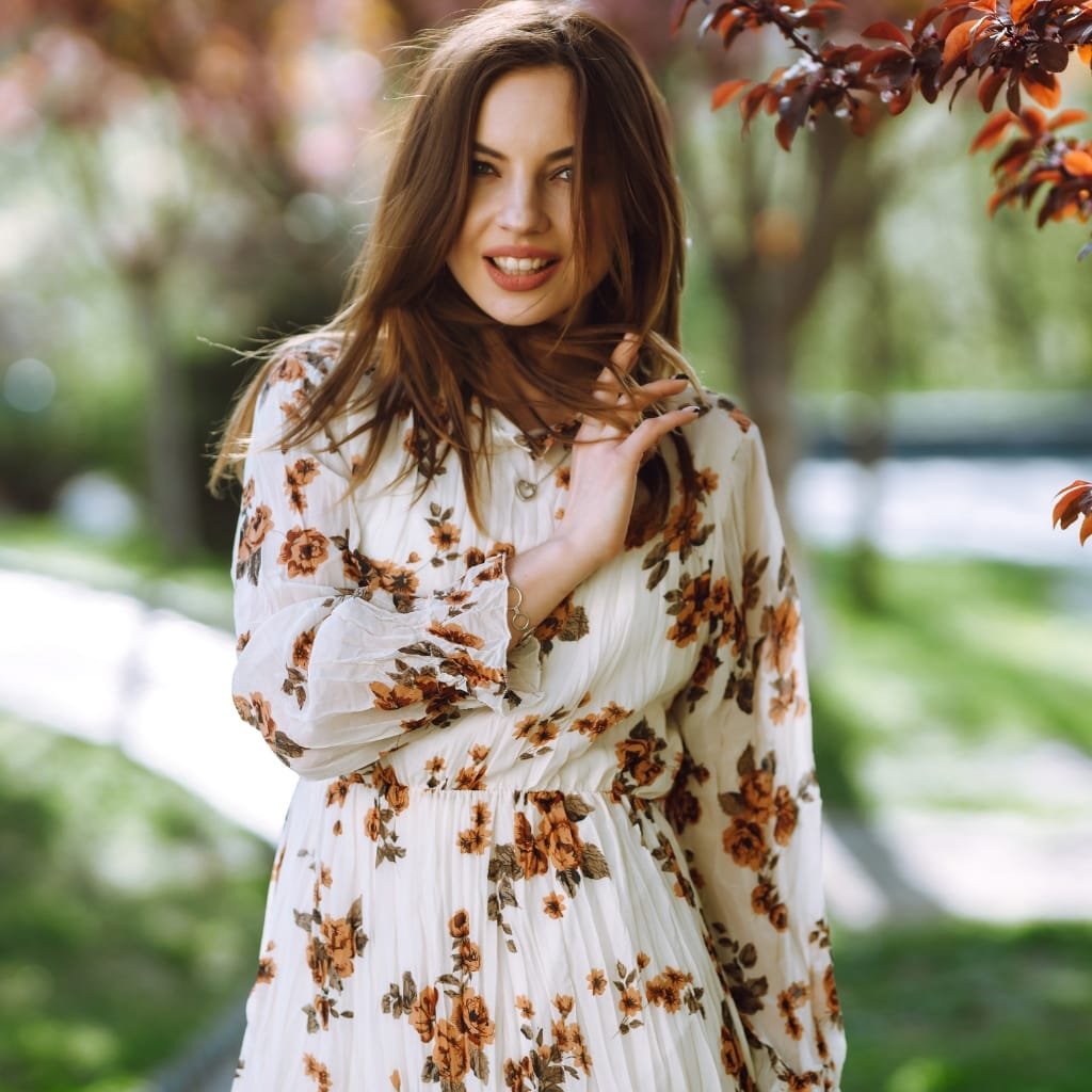A woman with long, wavy brown hair smiling playfully while standing outdoors near a tree with orange and red leaves. She wears a long-sleeved, white chiffon dress with a floral pattern in shades of brown and orange, suitable for transitional seasons.