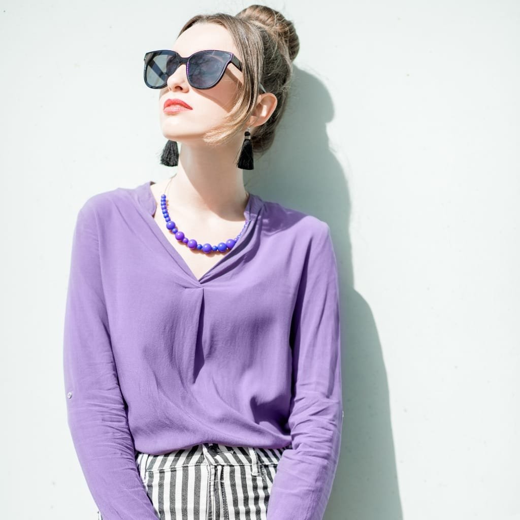 A woman wearing a light purple polyester chiffon blouse, paired with striped pants, accessorized with a blue beaded necklace and black tassel earrings, and sporting large sunglasses against a plain white wall.