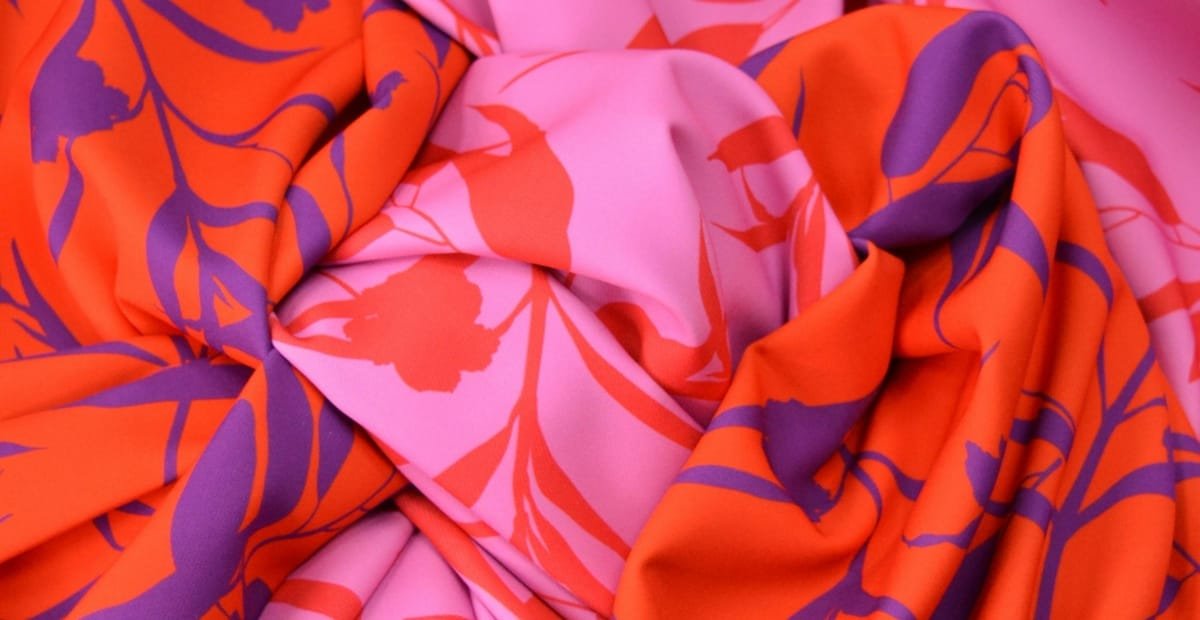 Close-up image of pink and orange chiffon fabric with purple floral or botanical prints, showing the soft, flowing texture and vibrant print design.