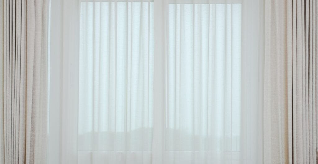 Sheer white curtains with beige drapes on either side, hanging in front of a window.
