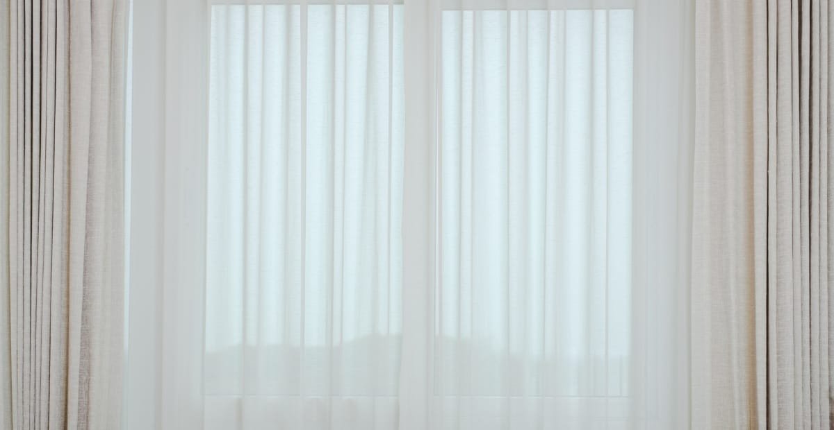 Sheer white curtains with beige drapes on either side, hanging in front of a window.