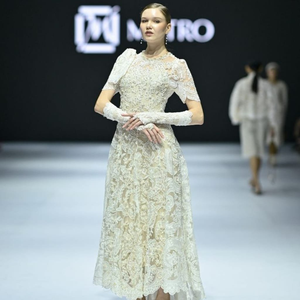A woman walking on a runway wearing an elegant, intricate cathedral lace dress with matching lace gloves. The background is dark with a logo and some people in the distance.