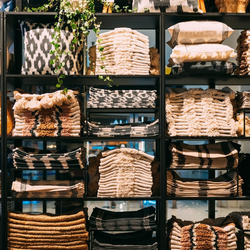 A store display featuring neatly stacked rolls of high-quality basket-weave cotton fabric in various neutral tones and patterns, arranged on black shelving units.
