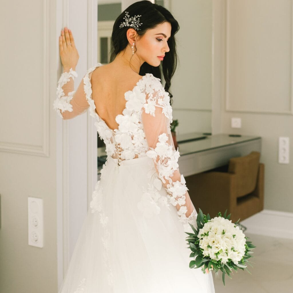 A bride wearing a beautiful white lace wedding dress with long sheer sleeves decorated with floral appliqués, an open back, and holding a bouquet of white flowers.