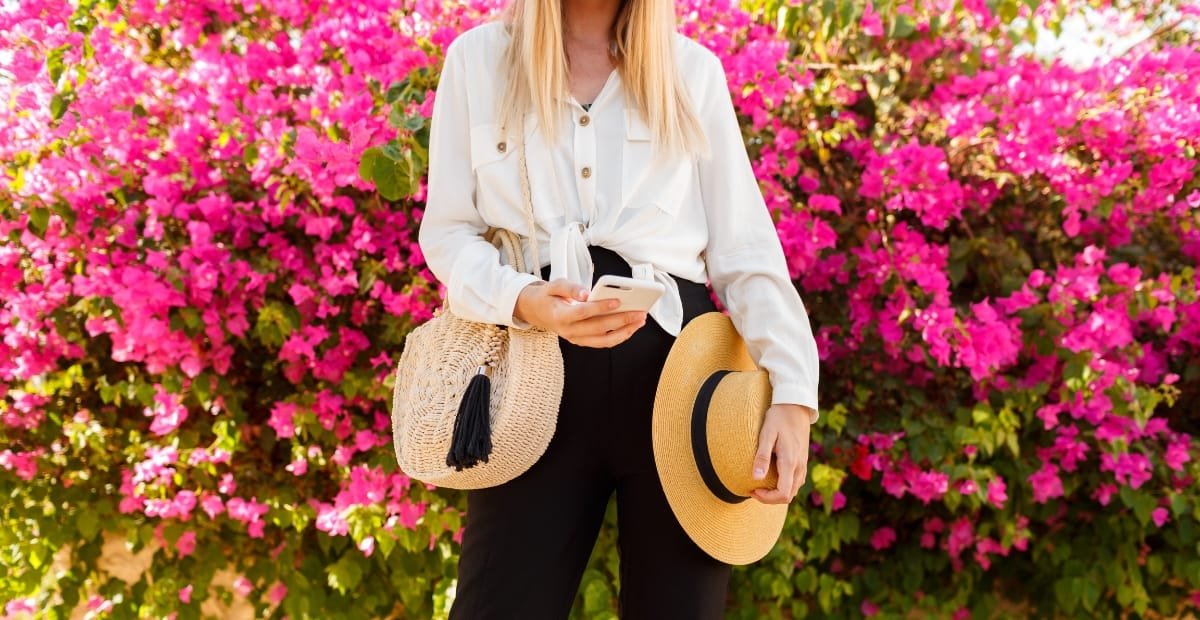 A woman holding a smartphone and a wide-brimmed straw hat, standing in front of vibrant pink flowering bushes, with a straw handbag hanging from her shoulder.
