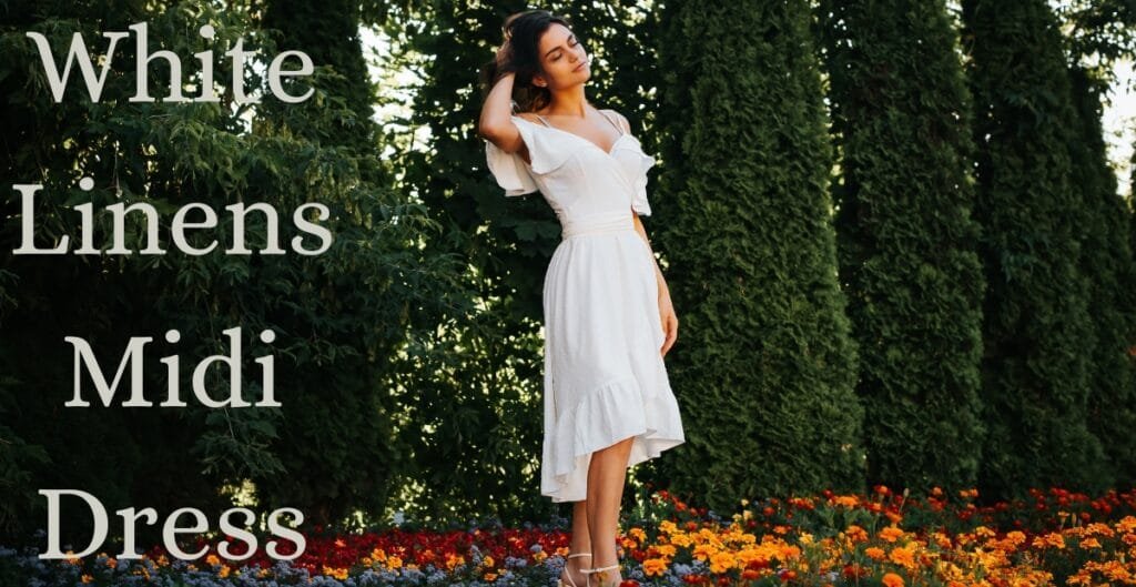 A woman wearing a white midi dress with ruffled sleeves standing outdoors among trees and colorful flowers, with sunlight filtering through the foliage.