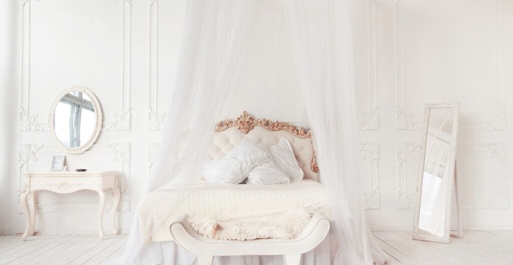 A luxurious bedroom with a white, ornate bed draped with sheer curtains, a matching white vanity table with a mirror, and a full-length mirror leaning against the wall. The room features intricate wall moldings and a soft, elegant atmosphere.
