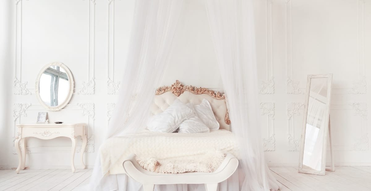 A luxurious bedroom with a white, ornate bed draped with sheer curtains, a matching white vanity table with a mirror, and a full-length mirror leaning against the wall. The room features intricate wall moldings and a soft, elegant atmosphere.