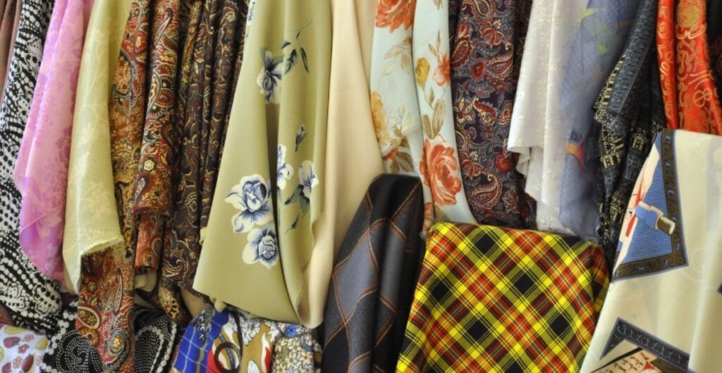 A collection of colorful fabrics hanging side by side, featuring various patterns including floral, paisley, checkered, and abstract designs.