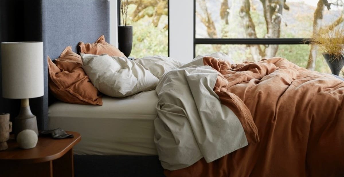 A cozy bedroom with an unmade bed featuring white and rust-colored bedding, a nightstand with a lamp, a book, and decorative objects, and large windows allowing natural light to brighten the room, with trees visible outside.
