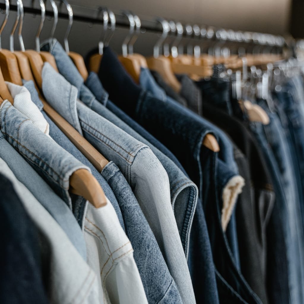 Close-up of a row of chambray shirts hanging on wooden hangers on a clothing rack, showing the fabric's texture and color.