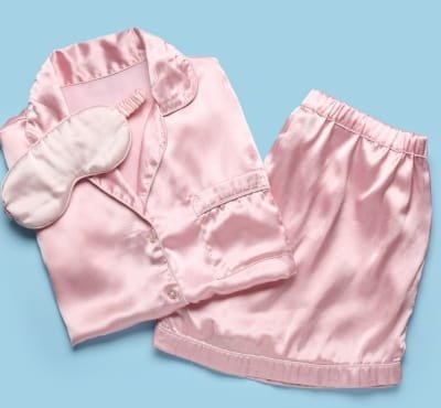 A set of pink satin pajamas including a long-sleeve shirt, shorts, and slippers, laid out on a light blue background.