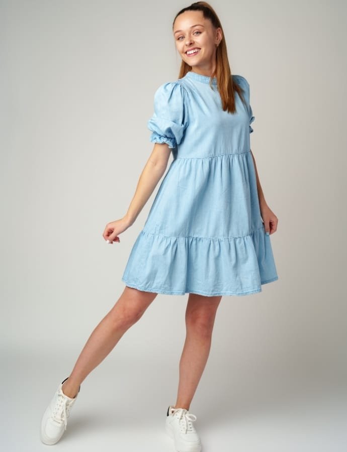 Young woman smiling and posing in a light blue smock cotton dress, paired with white sneakers, against a neutral background.