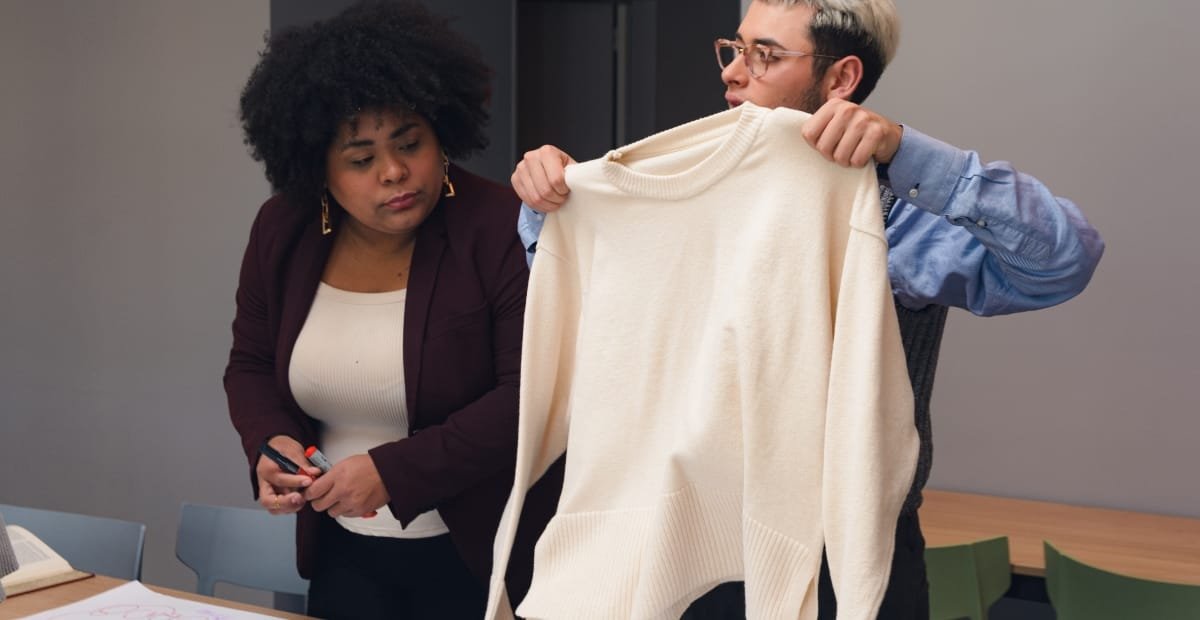 A man and a woman are in an office, with the man holding up a large, cream-colored sweater while the woman looks on attentively, holding a pen.