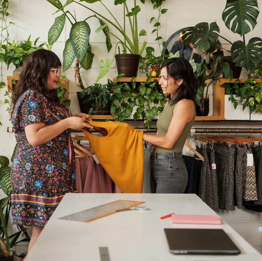 Two women are shopping for clothing in a boutique filled with green plants and wooden shelves, with one woman showing a mustard-yellow fabric to the other.