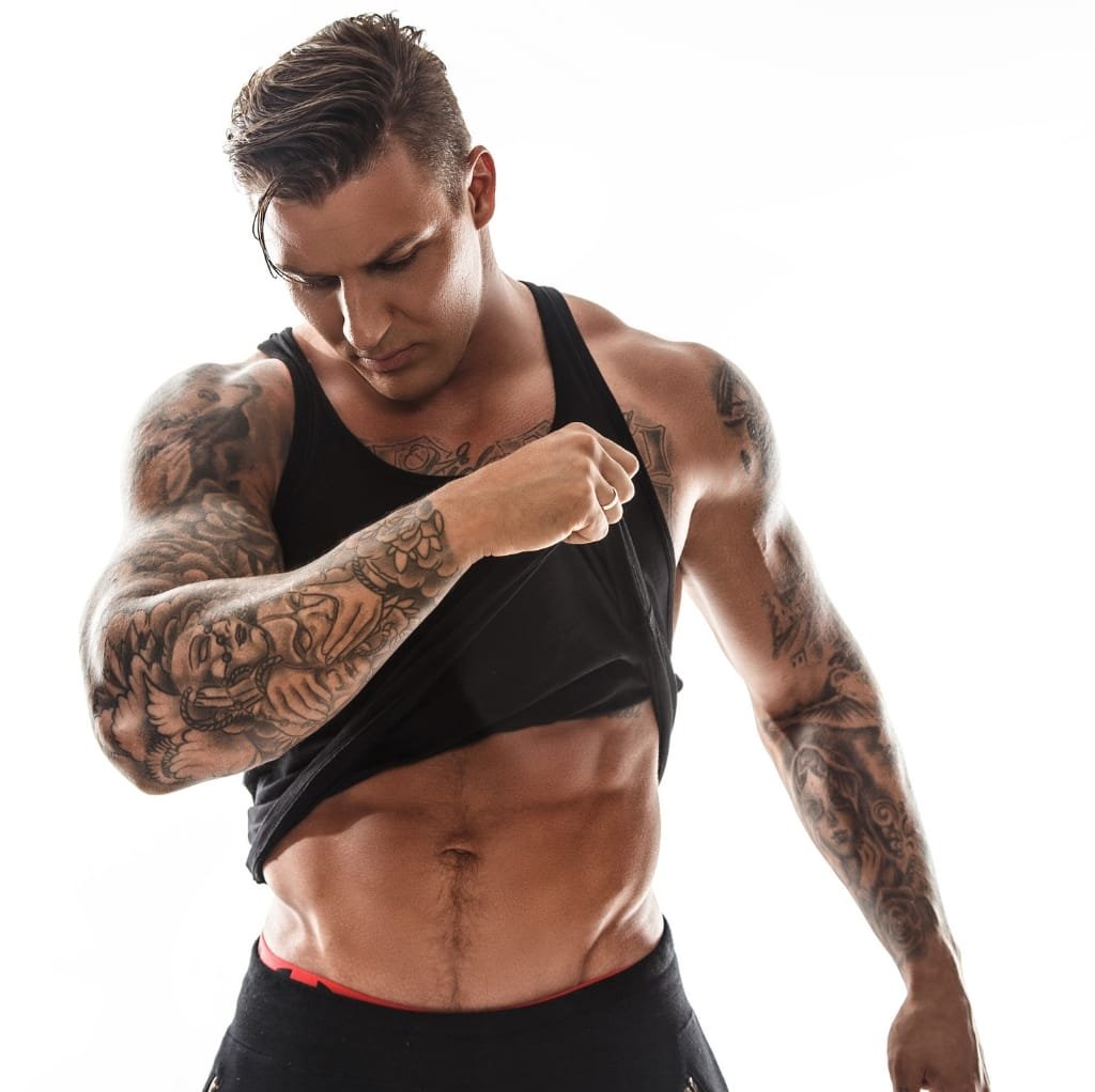 A muscular man with tattoos lifting his black sports tank top to reveal defined abs and chest muscles, standing against a plain white background.