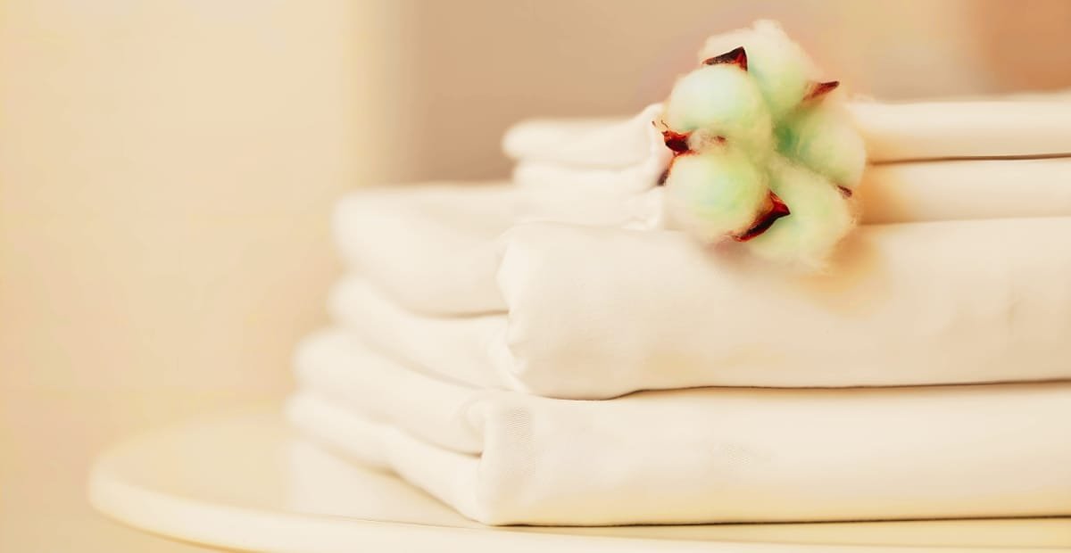 A stack of neatly folded silk sheets with a small cluster of cotton bolls resting on top, against a soft, neutral background.