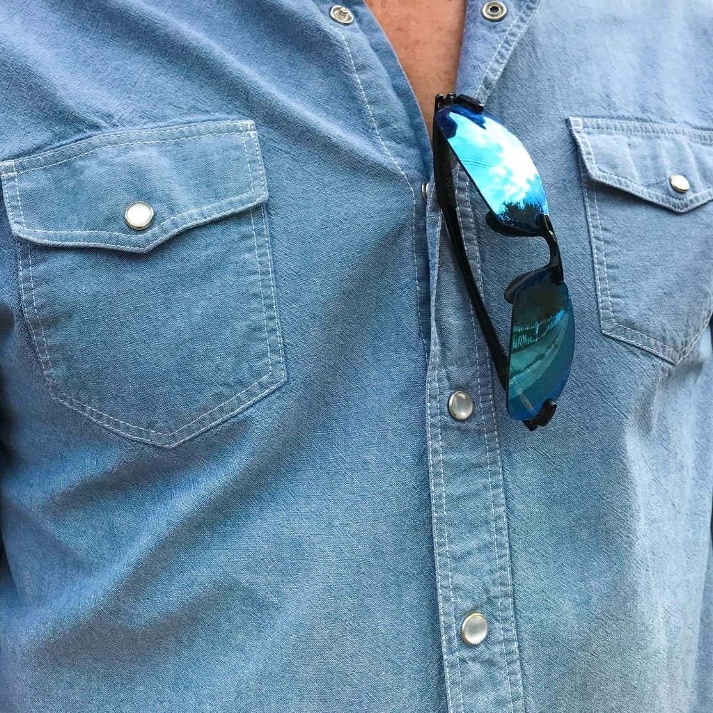Close-up of a denim-like chambray shirt with chest pockets, featuring a pair of reflective sunglasses clipped onto the collar.