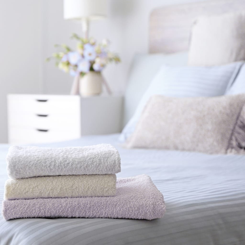 A neatly folded stack of three towels on a bed, with a bedroom in the background featuring a bedside table, a vase with flowers, and pillows.